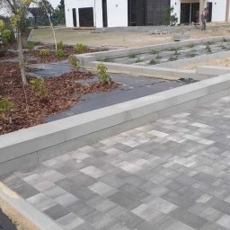 Brukarstwo Babiarz - Ułożona szara kostka brukowa na podjeździe w kształcie litery L, z obrzeżami z betonowych elementów, widoczne przygotowane rabaty z agrowłókniną i korą, w tle dom jednorodzinny i pracownik.
