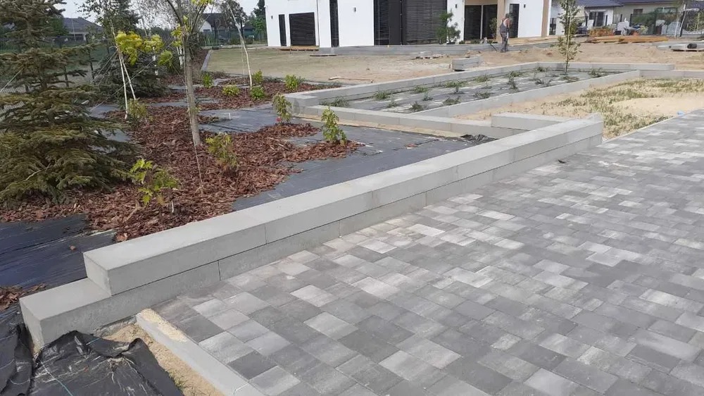 Ułożona szara kostka brukowa na podjeździe w kształcie litery L, z obrzeżami z betonowych elementów, widoczne przygotowane rabaty z agrowłókniną i korą, w tle dom jednorodzinny i pracownik.