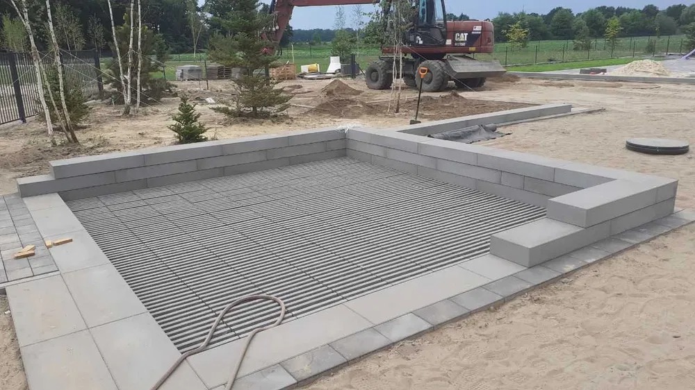 Budowa basenu: Szary szkielet basenu z kratką na dnie, otoczony betonowymi blokami i kostką brukową, z koparką CAT w tle na piaszczystym terenie.