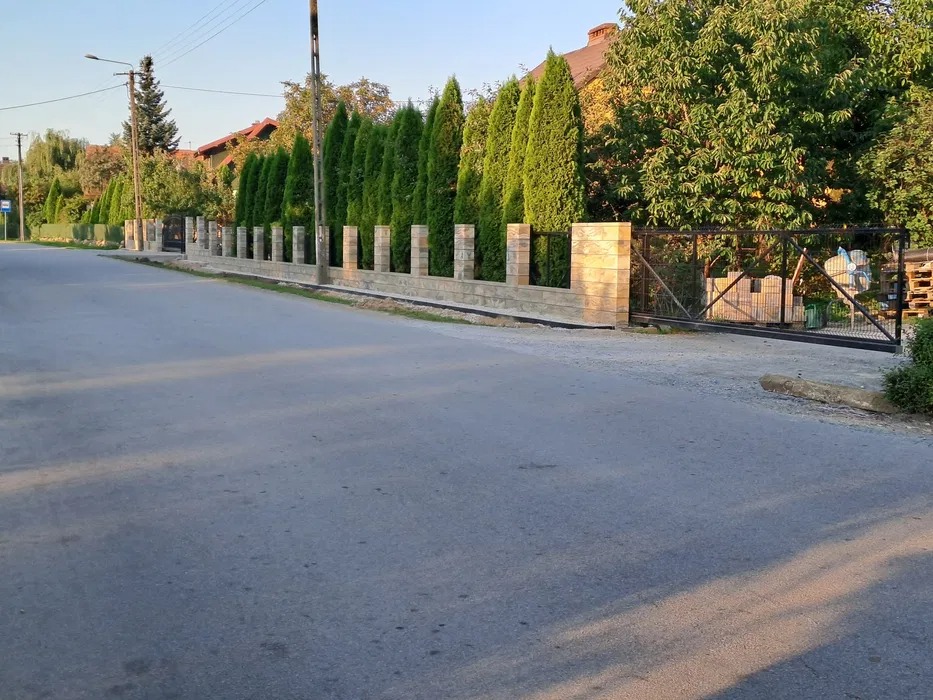 Nowoczesne ogrodzenie z jasnych bloczków z betonowymi słupkami i metalową bramą wjazdową, przylegające do asfaltowej drogi i szpaleru wysokich tuj.