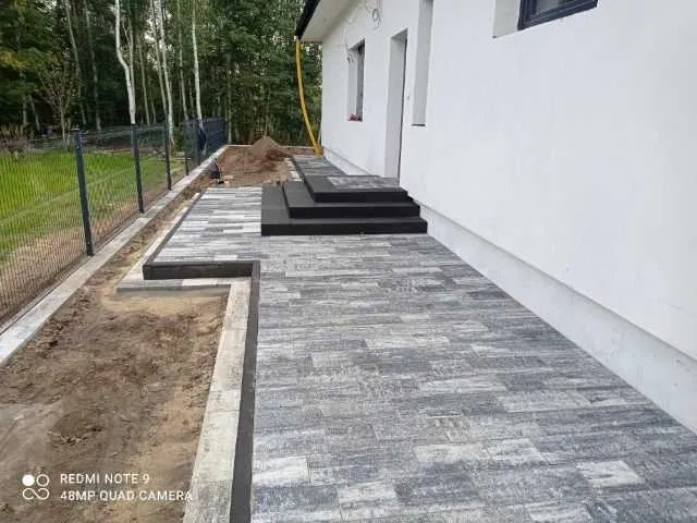 Ułożona szara kostka brukowa przed białym budynkiem z czarnymi schodami, widoczny fragment ogrodzenia i przygotowane podłoże z ziemią.
