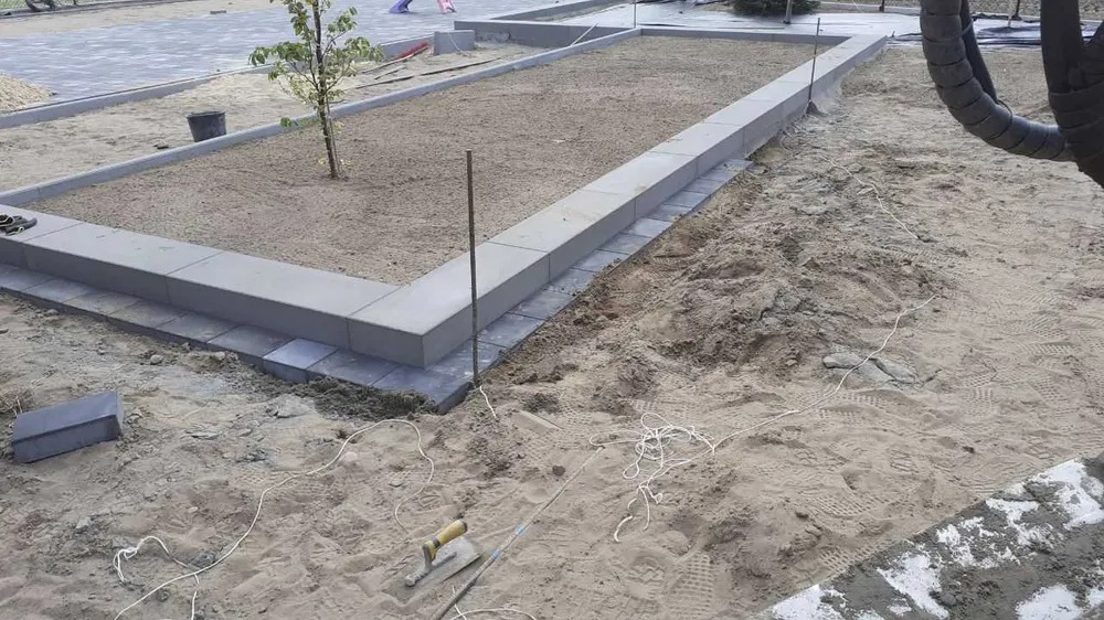 Przygotowanie terenu pod trawnik lub ogród: obrzeża z szarej kostki betonowej, wyrównana powierzchnia piasku, sznurek wyznaczający linie, kielnia na piasku.