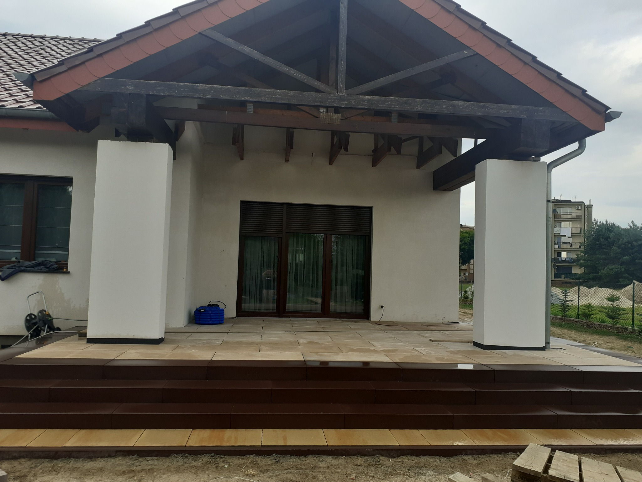 Wyłożony kostką brukową taras z brązowymi schodami przed domem w Oleśnicy, widoczne białe filary podtrzymujące drewnianą konstrukcję dachu.