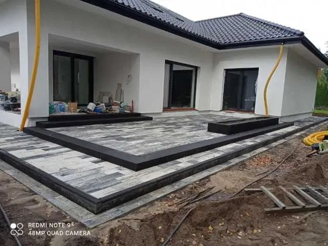 Ułożona kostka brukowa przed nowoczesnym domem z czarną stolarką okienną i żółtą rurą spustową, widoczne obrzeża i przygotowany teren pod dalsze prace.