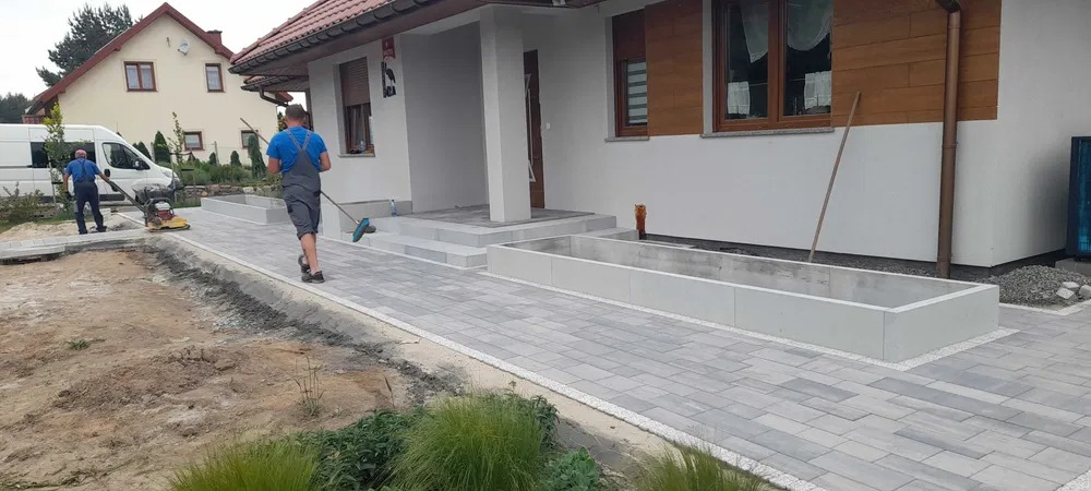 Pracownicy układają szarą kostkę brukową przed domem jednorodzinnym z białymi donicami, jeden zamiata, drugi ubija teren zagęszczarką, widoczny bus firmy.