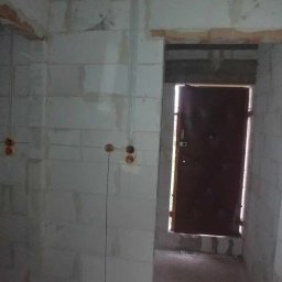 Marcin Owsiany - Surowy stan wnętrza z instalacją elektryczną: kable w peszlach prowadzone po ścianie z bloczków, widoczne puszki pod gniazdka, betonowy strop i drzwi wejściowe.