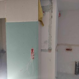 Marcin Owsiany - Wnętrze w trakcie remontu: widoczne przewody elektryczne wystające z puszki w płycie kartonowo-gipsowej, kable prowadzone w ścianie, częściowo zakryte żółtą tkaniną, ściany w stanie surowym.