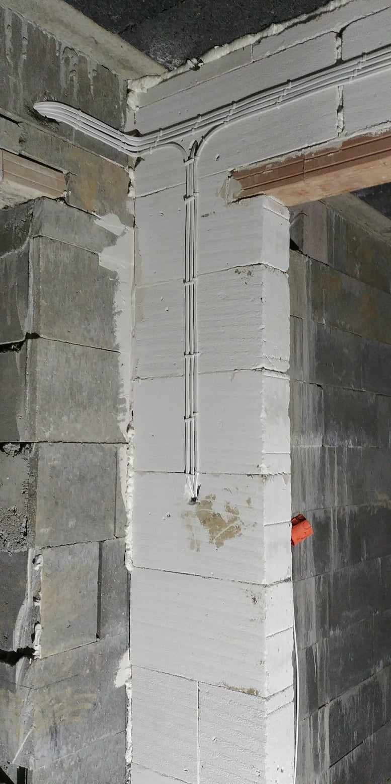 Kable elektryczne prowadzone po ścianie z bloczków betonowych, zabezpieczone uchwytami, widoczna puszka instalacyjna w kolorze pomarańczowym.