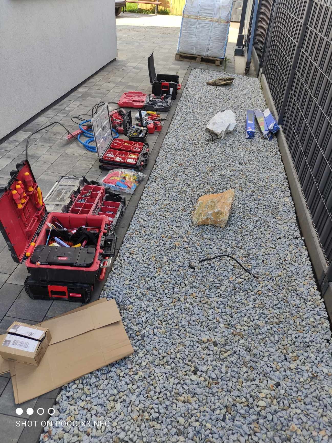 Rozłożone skrzynki narzędziowe z osprzętem elektrycznym na kostce brukowej i żwirowym podłożu wzdłuż ogrodzenia.
