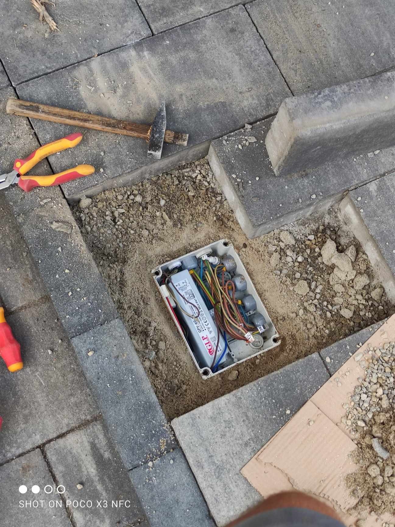 Otwarta puszka elektryczna z przewodami i zasilaczem LED, umieszczona w wykopie w kostce brukowej, obok leżą młotek, kombinerki i fragment kartonu.