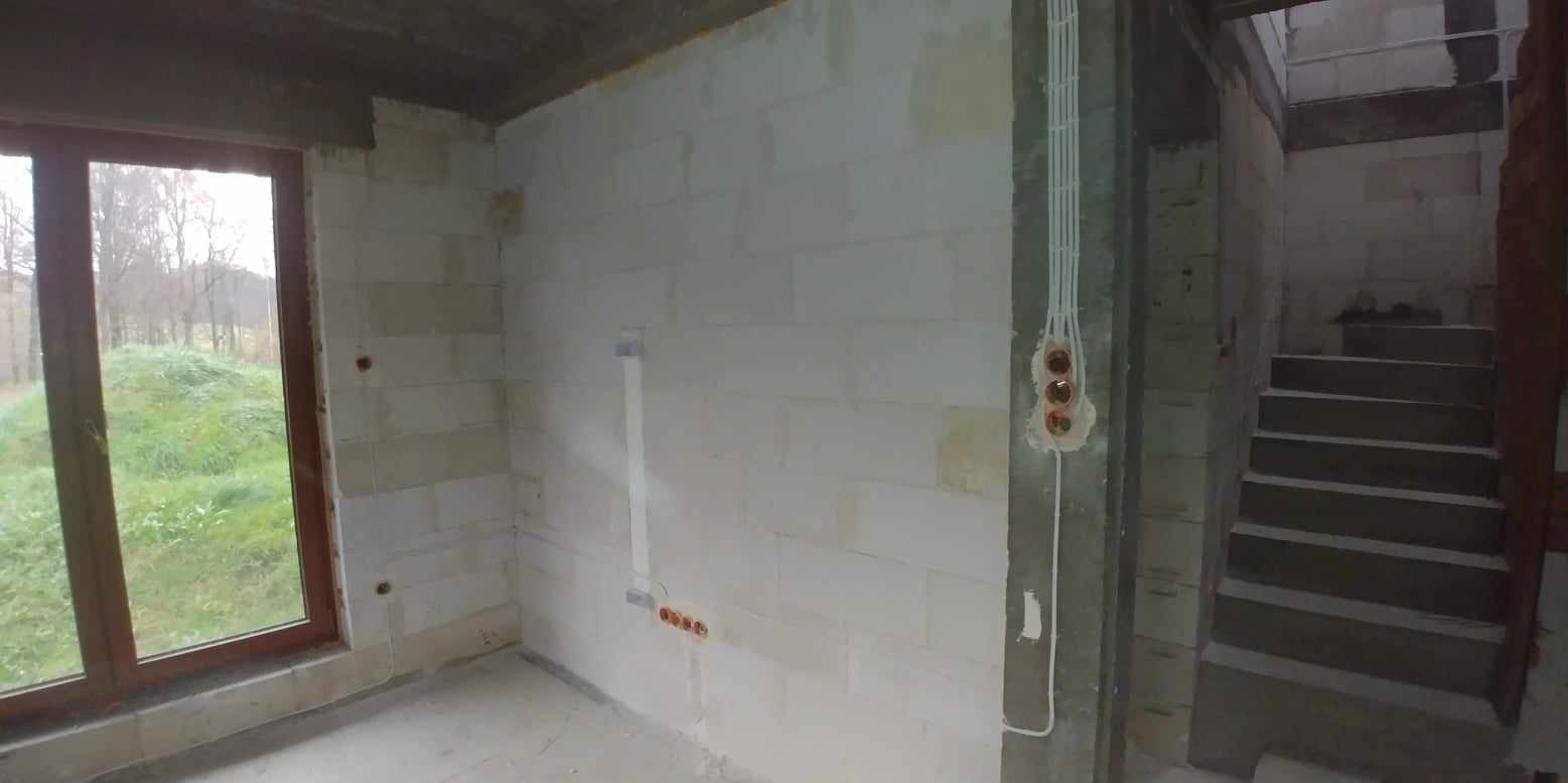 Surowy stan wnętrza domu w trakcie instalacji elektrycznej: widoczne przewody w ścianie, puszki instalacyjne i okno z widokiem na zewnątrz.