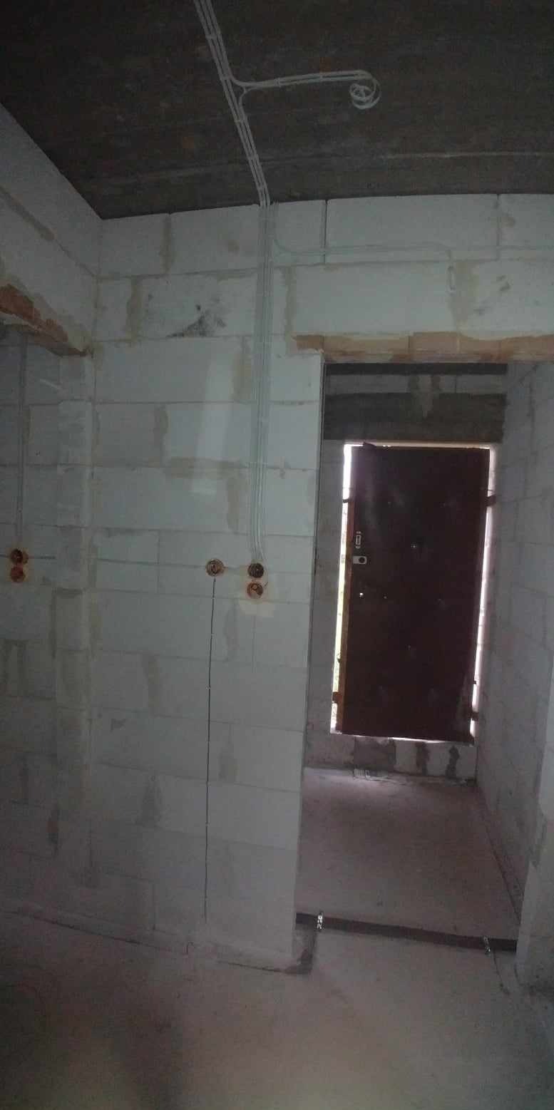 Surowy stan wnętrza z instalacją elektryczną: kable w peszlach prowadzone po ścianie z bloczków, widoczne puszki pod gniazdka, betonowy strop i drzwi wejściowe.
