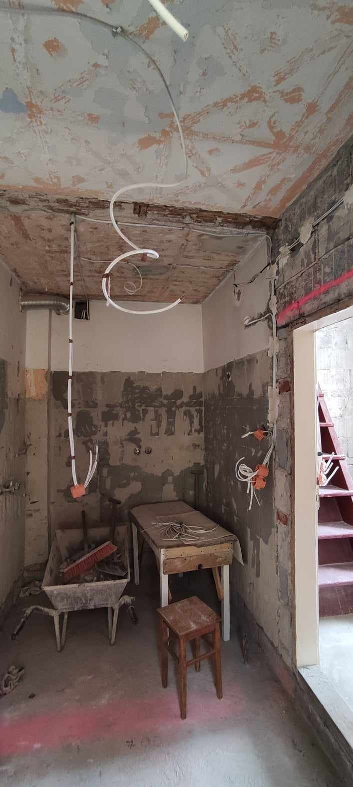 Wnętrze remontowanego pomieszczenia z widocznymi przewodami elektrycznymi, puszkami instalacyjnymi, taczką z narzędziami i drewnianym stołem, na ścianach ślady po skuciu tynku, surowy stan...