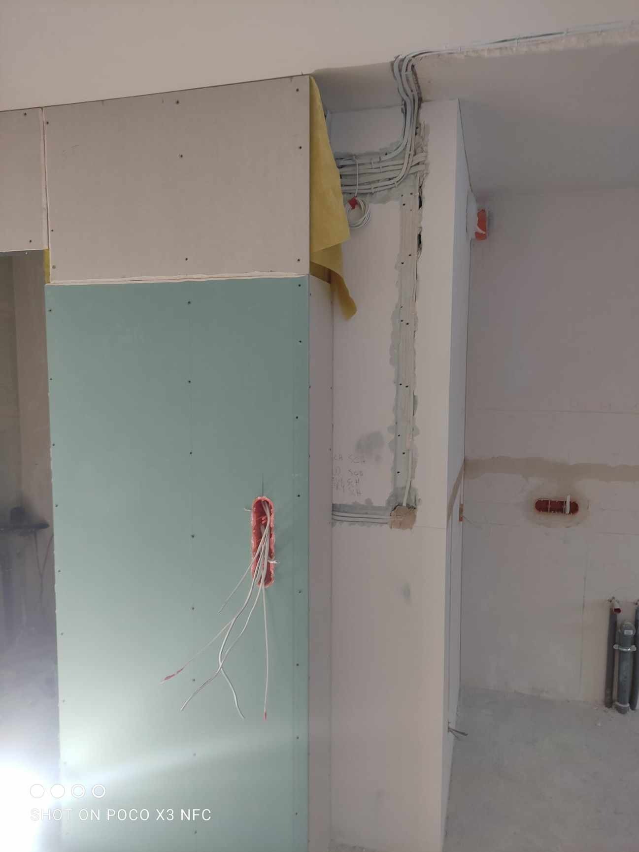Wnętrze w trakcie remontu: widoczne przewody elektryczne wystające z puszki w płycie kartonowo-gipsowej, kable prowadzone w ścianie, częściowo zakryte żółtą tkaniną, ściany w stanie surowym.