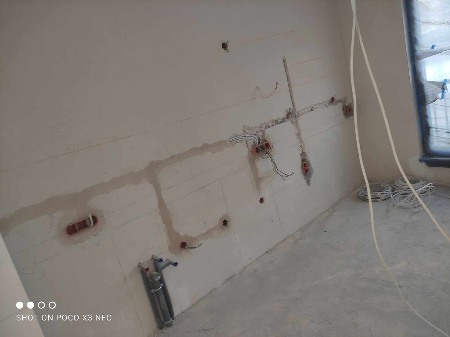 Ściana w trakcie remontu z widocznymi przewodami i puszkami elektrycznymi oraz rurami kanalizacyjnymi, szpachlowane bruzdy, kable zwisające ze ściany.