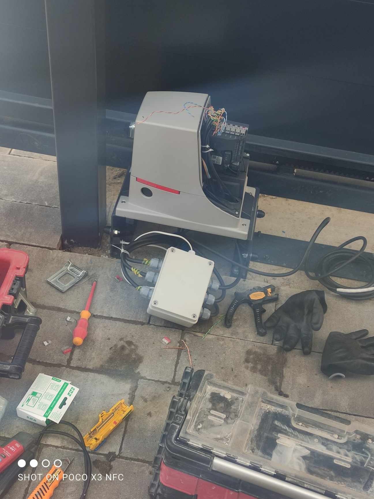 Montaż automatyki bramy przesuwnej: widoczny mechanizm z podłączonymi przewodami, skrzynka sterownicza, narzędzia (śrubokręt, obcinak do kabli), rękawice robocze i skrzynka narzędziowa...