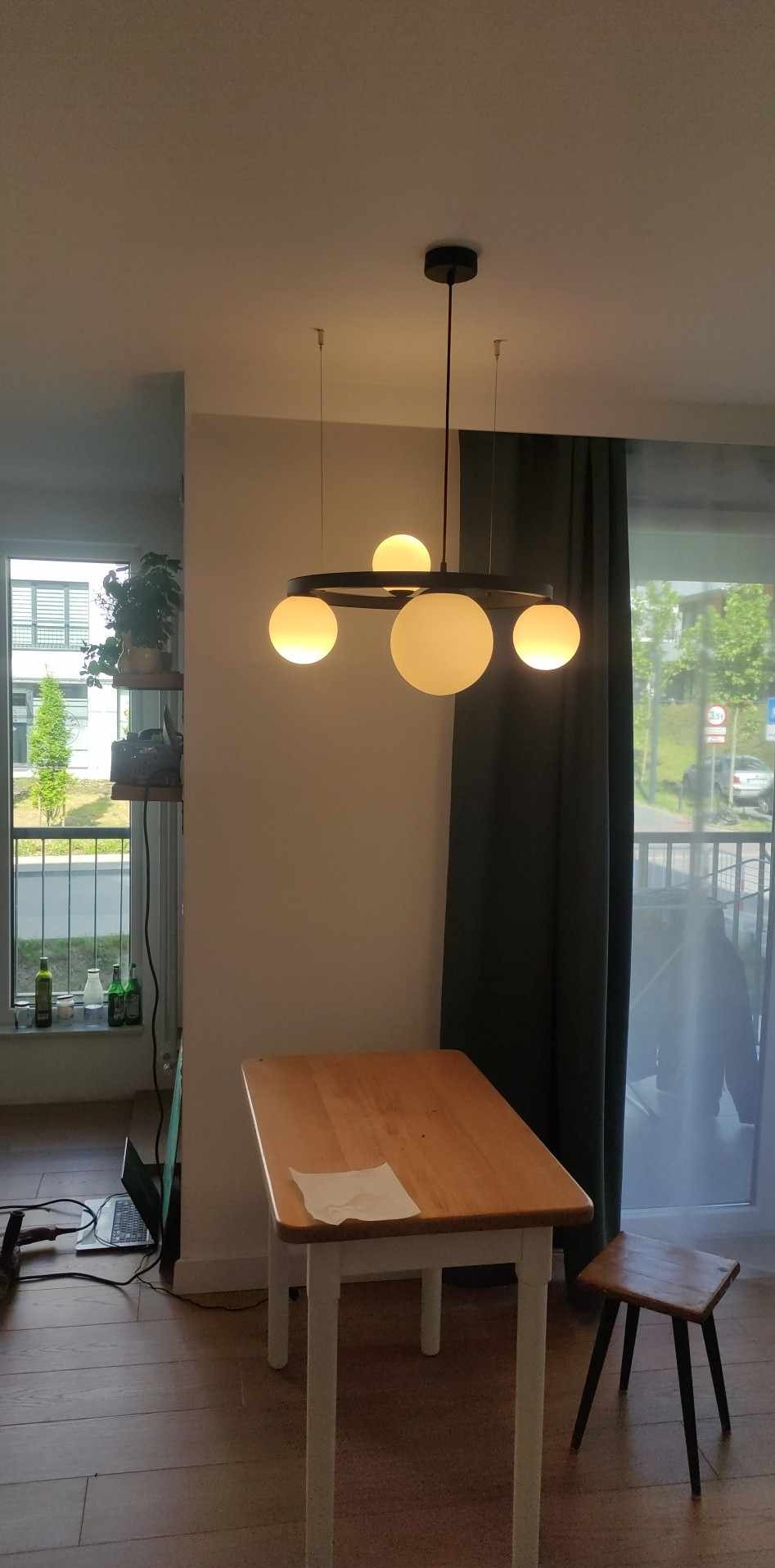 Nowoczesna lampa wisząca z okrągłą obręczą i pięcioma szklanymi kulami w jasnym wnętrzu, widok na okno z zielenią i stolik z krzesłem w tle.