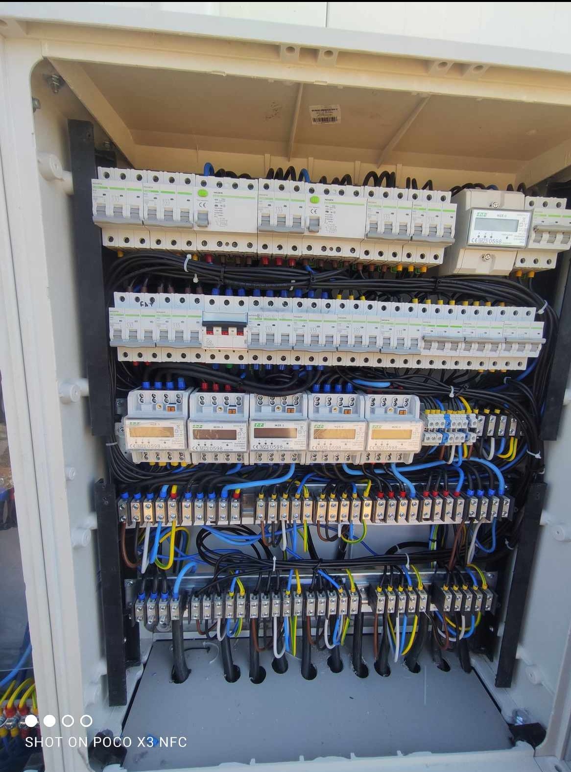 Otwarta skrzynka elektryczna z zamontowanymi bezpiecznikami, licznikami energii i okablowaniem w kolorach: czarnym, niebieskim, żółtym i brązowym.