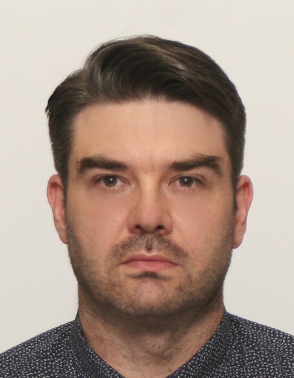 Portret mężczyzny z krótkimi, ciemnymi włosami i lekkim zarostem, ubrany w koszulę w drobny wzór, na jasnym tle. Ujęcie od przodu, skupione na twarzy.