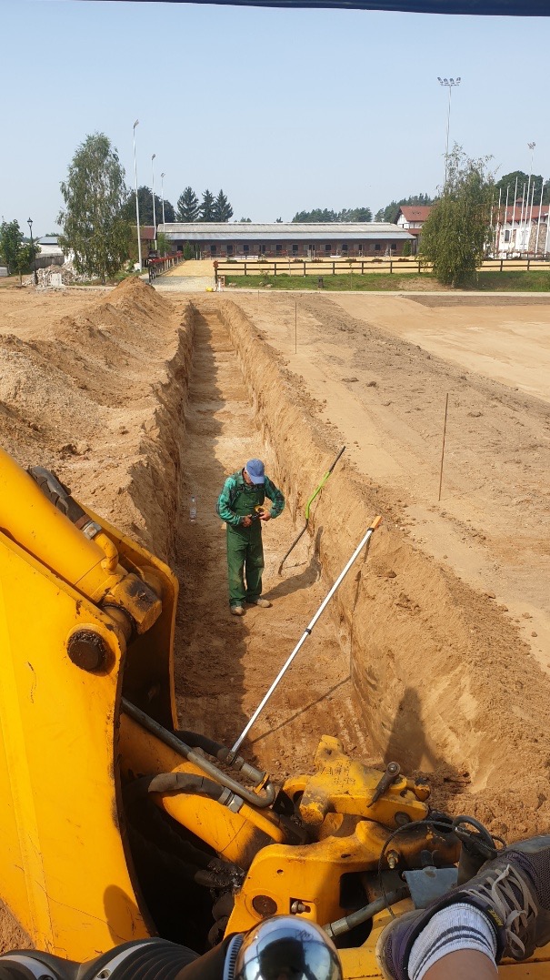 Wąski wykop w piaszczystej ziemi, pracownik w kombinezonie mierzy głębokość, widoczny fragment żółtej koparki i budynki w tle.