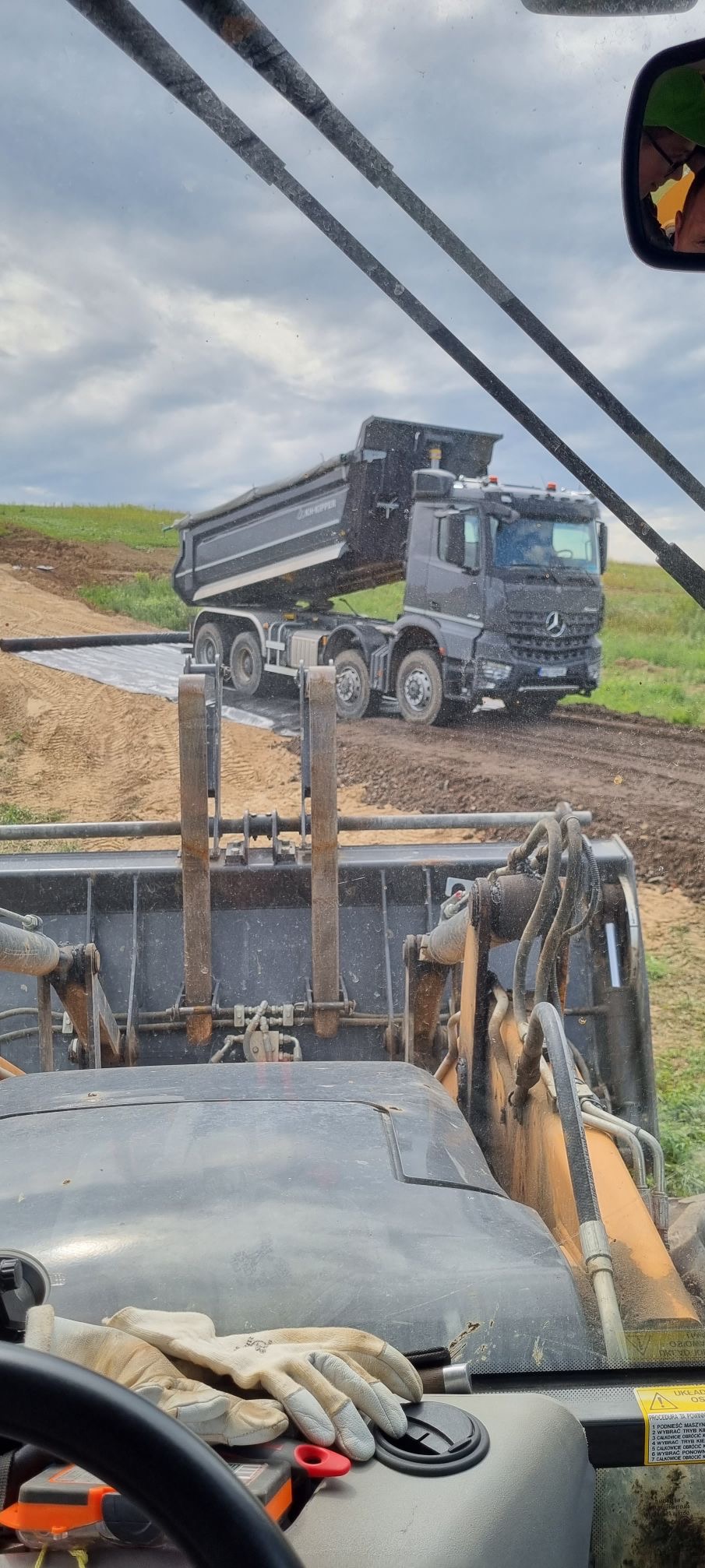 Widok z kabiny ładowarki teleskopowej na wywrotkę Mercedes-Benz podczas rozładunku ziemi na rozłożoną geowłókninę na polu, pochmurne niebo w tle.