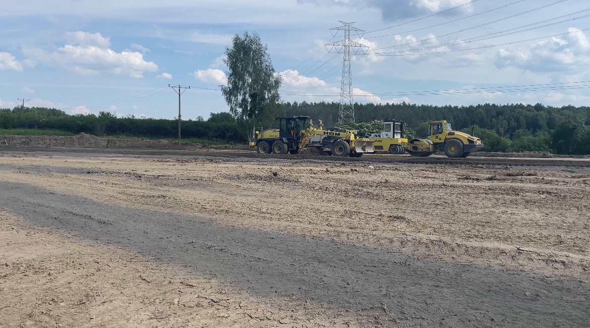 Żółte walce drogowe i równiarka HBM pracują na rozległym, przygotowanym terenie budowlanym w słoneczny dzień, z widoczną linią wysokiego napięcia w tle.