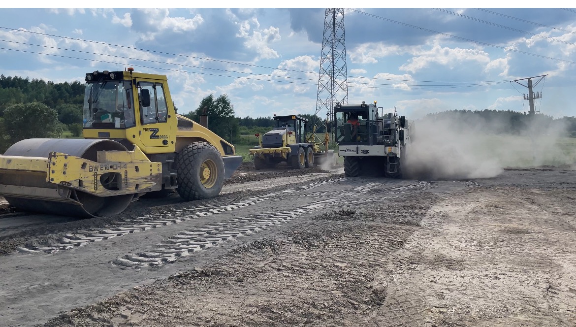 Żółty walec drogowy Bomag BW 216-DH-4 ubija ziemię na placu budowy, w tle pracują inne maszyny budowlane i widać słupy wysokiego napięcia.
