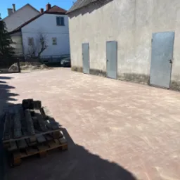 Nowo ułożona, czerwona kostka brukowa na podwórku z paletą kostki w rogu, obok budynku z trzema metalowymi drzwiami i domu w tle.