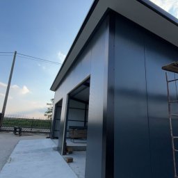 Patryk Kraszewski - Niedokończona budowa garażu z czarnych paneli, widoczne elementy konstrukcyjne i drabina, betonowa wylewka przed budynkiem, błękitne niebo w tle.