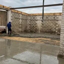 Wylewanie i rozprowadzanie betonu wewnątrz budynku z użyciem pompy i listwy wyrównującej, widoczne murowane ściany i drewniana konstrukcja stropu.