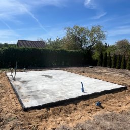 Patryk Kraszewski - Wybetonowana płyta fundamentowa na gruncie, z wystającymi rurami instalacyjnymi. W tle zieleń drzew i krzewów pod błękitnym niebem.