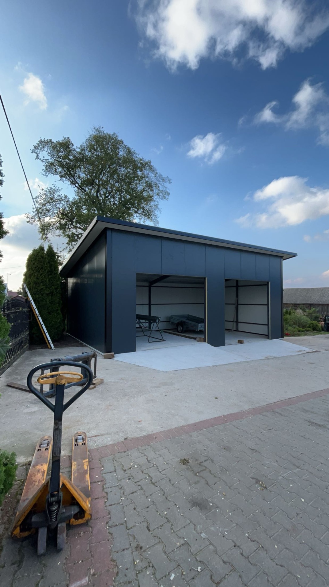 Nowoczesny, dwustanowiskowy garaż blaszany w kolorze grafitowym, z betonową wylewką, widziany z zewnątrz na tle błękitnego nieba z chmurami. Wewnątrz widoczne zaparkowane pojazdy i sprzęt. Obok...