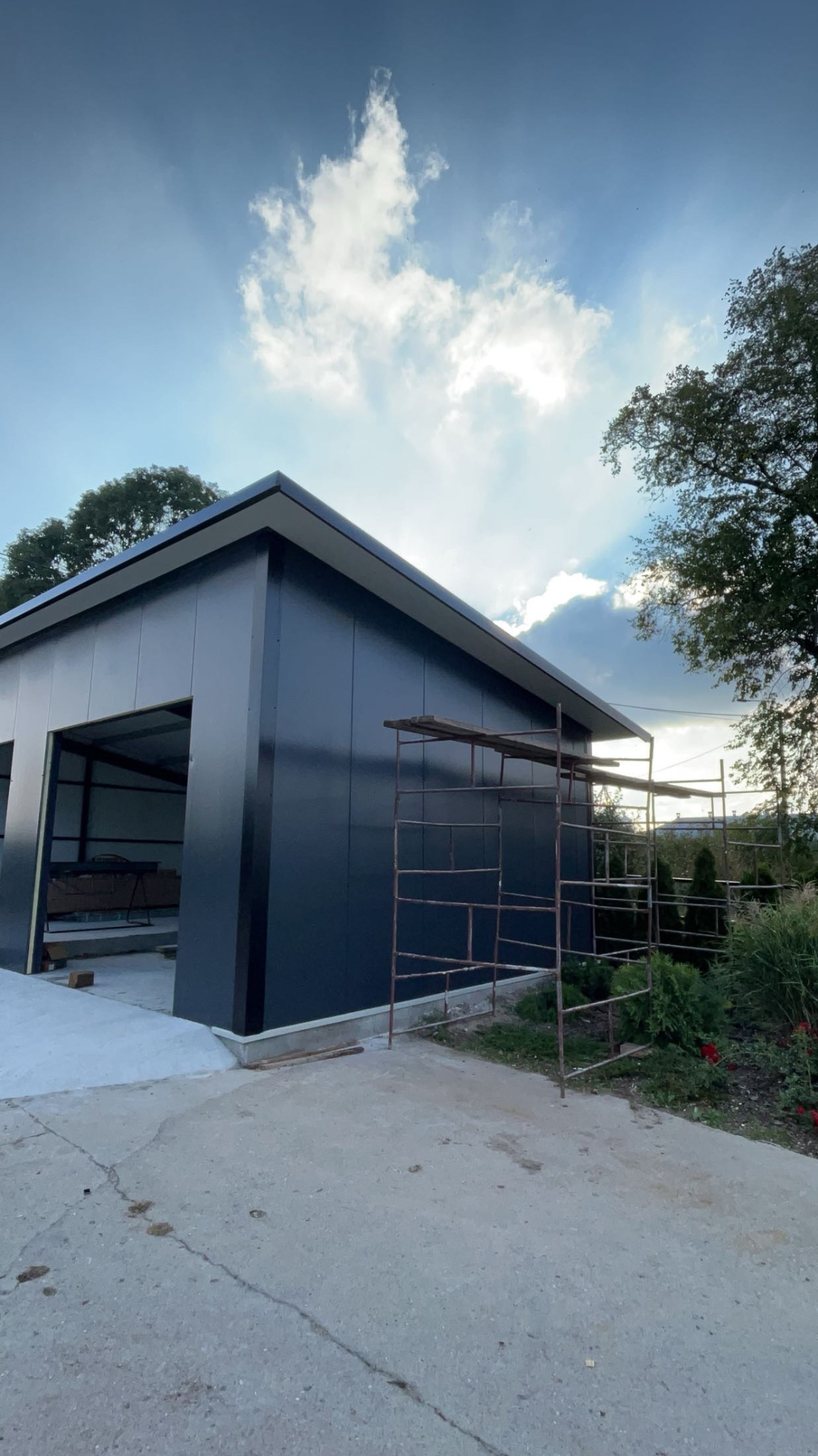 Nowoczesny, ciemny budynek gospodarczy z płaskim dachem w trakcie budowy, widoczne rusztowanie przy ścianie i częściowo otwarte wrota.