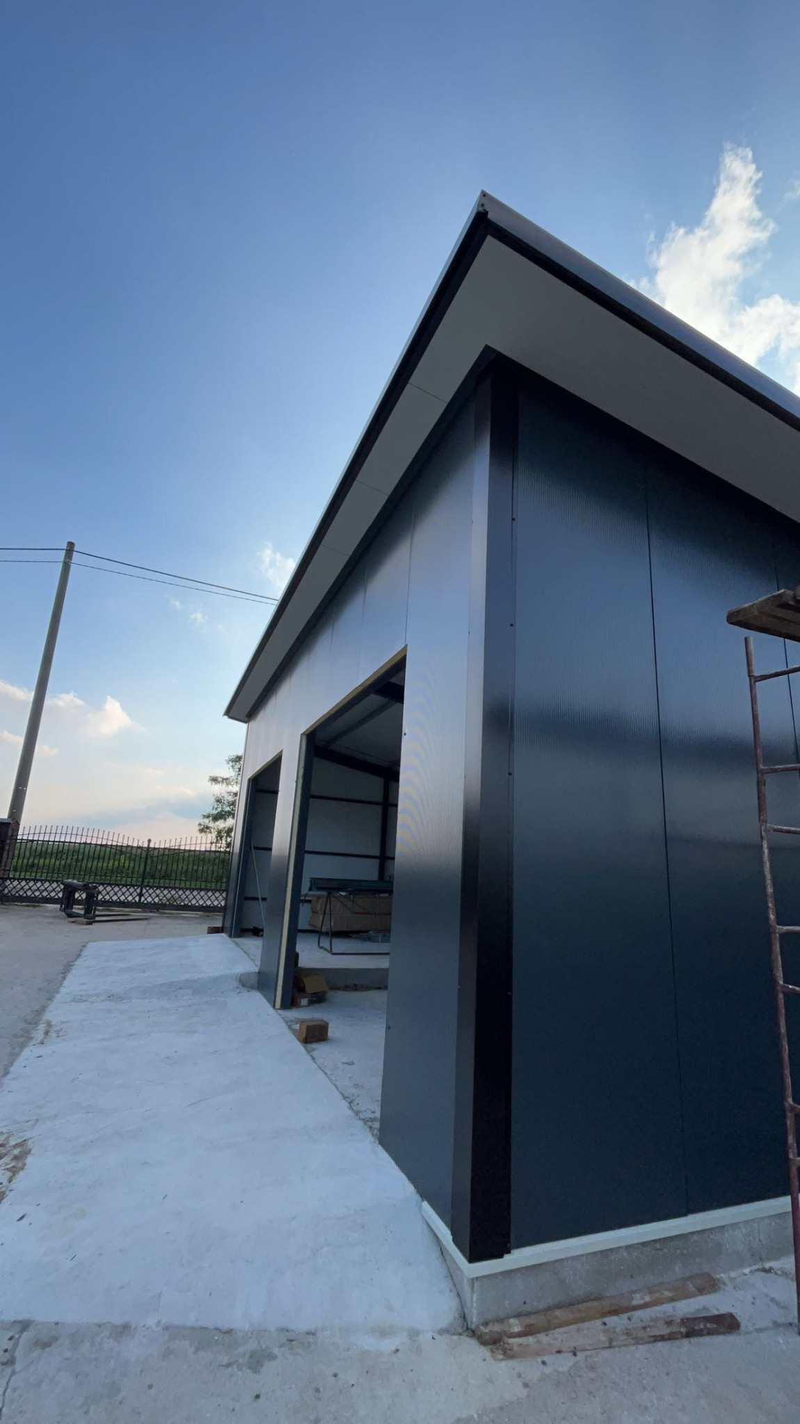 Niedokończona budowa garażu z czarnych paneli, widoczne elementy konstrukcyjne i drabina, betonowa wylewka przed budynkiem, błękitne niebo w tle.