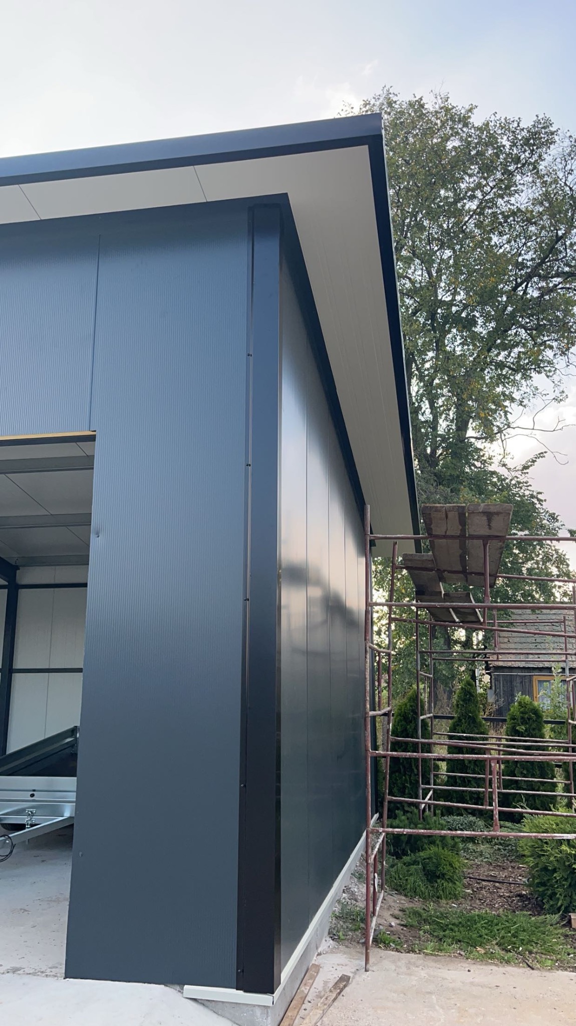 Nowoczesna, szara konstrukcja budynku z blachy trapezowej, widoczna z zewnątrz, z fragmentem rusztowania i zielenią w tle. Budynek w trakcie rozbudowy, widoczny fragment przyczepki.