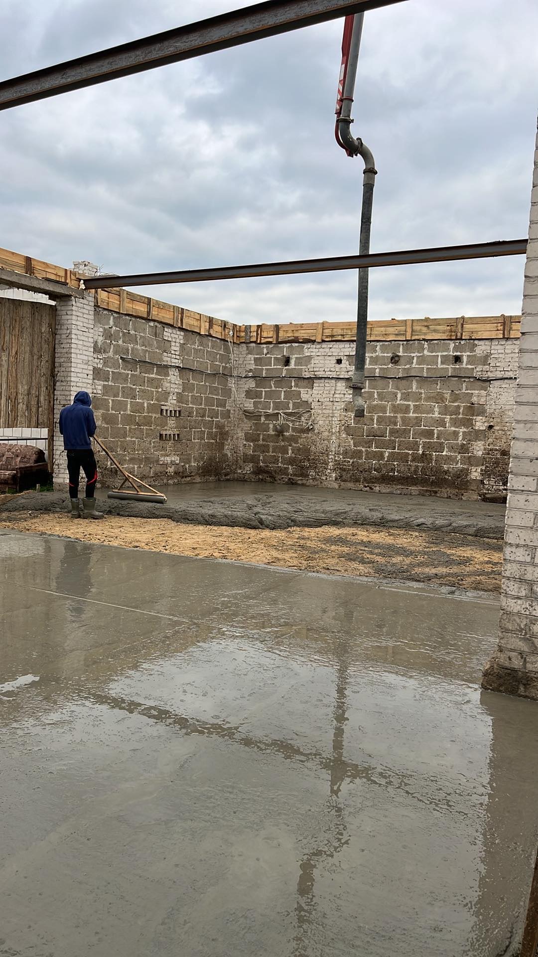 Wylewanie i rozprowadzanie betonu wewnątrz budynku z użyciem pompy i listwy wyrównującej, widoczne murowane ściany i drewniana konstrukcja stropu.