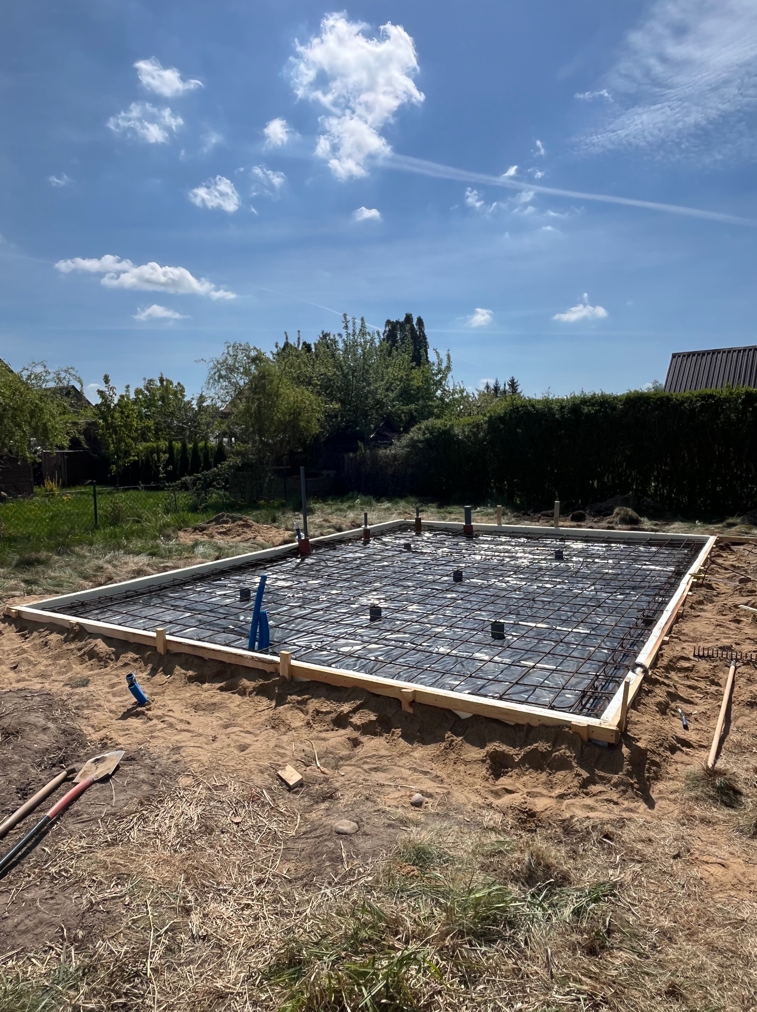 Przygotowany teren pod wylewkę fundamentów z deskowaniem, zbrojeniem i folią. Widoczne narzędzia pracy i fragment zabudowy w tle. Słoneczny dzień.