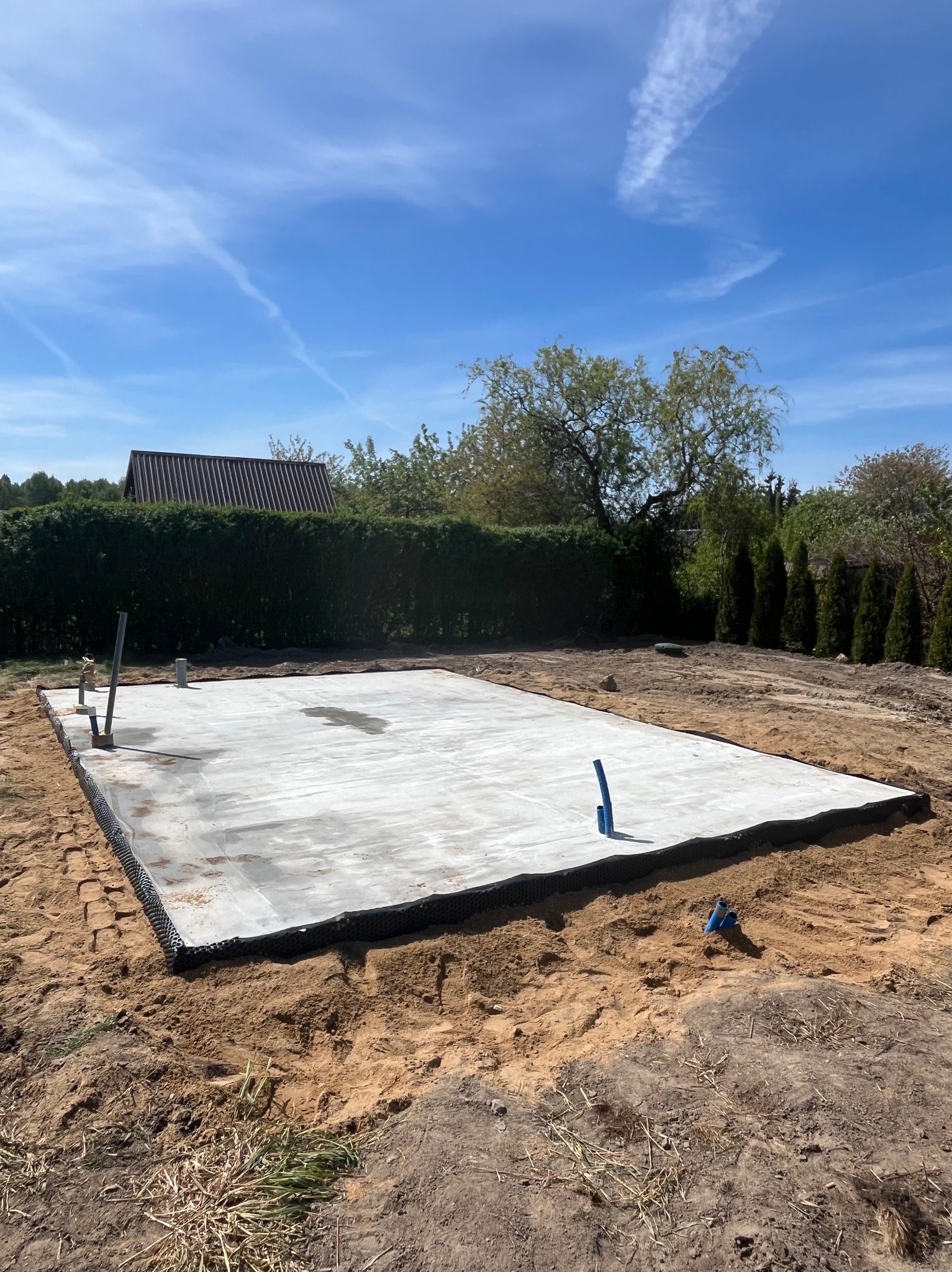 Wybetonowana płyta fundamentowa na gruncie, z wystającymi rurami instalacyjnymi. W tle zieleń drzew i krzewów pod błękitnym niebem.