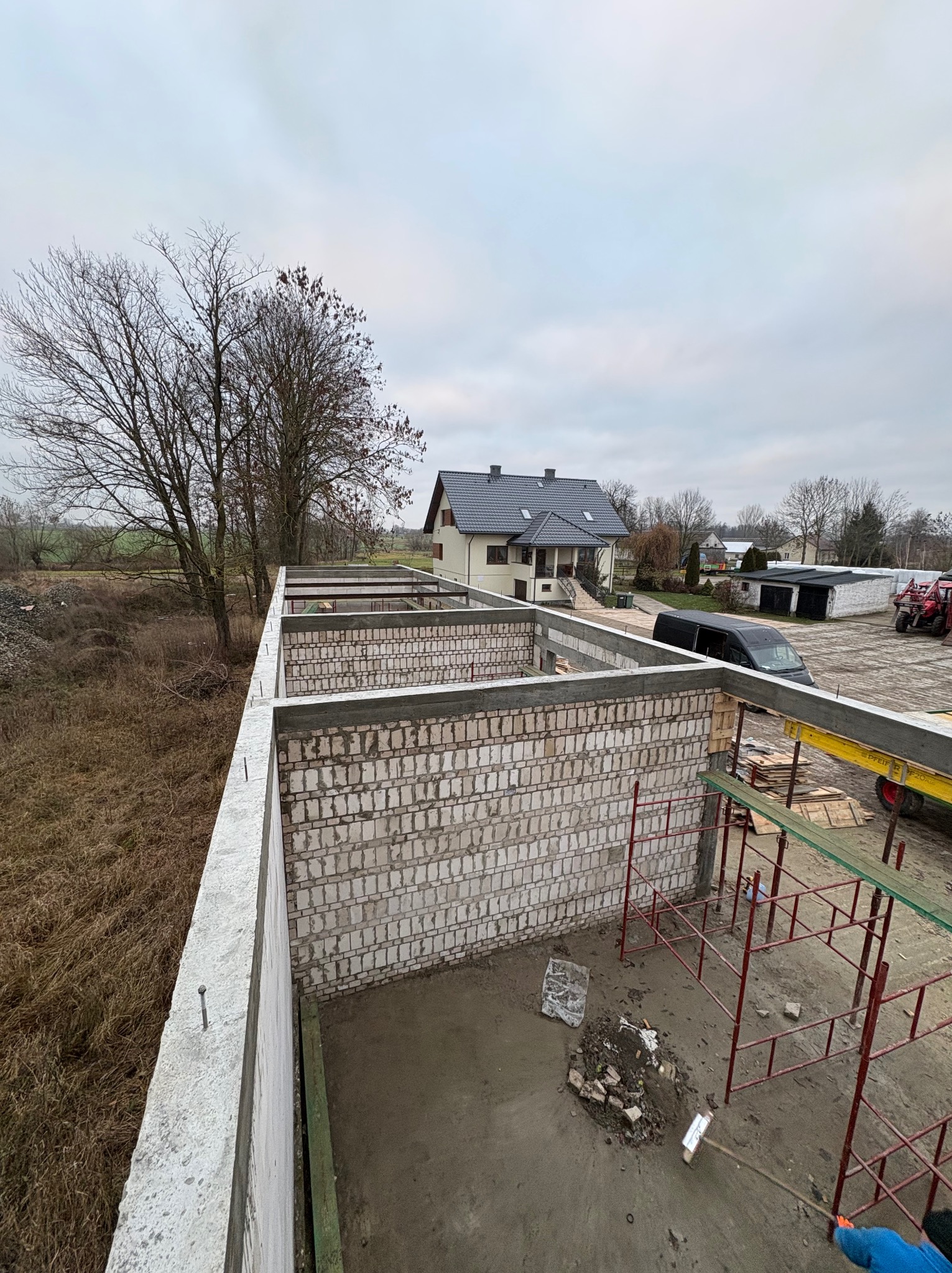 Wznoszone ściany z betonu komórkowego na budowie domu jednorodzinnego. Widoczne rusztowania, teren budowy i dom w tle. Ujęcie z góry, pochmurny dzień.