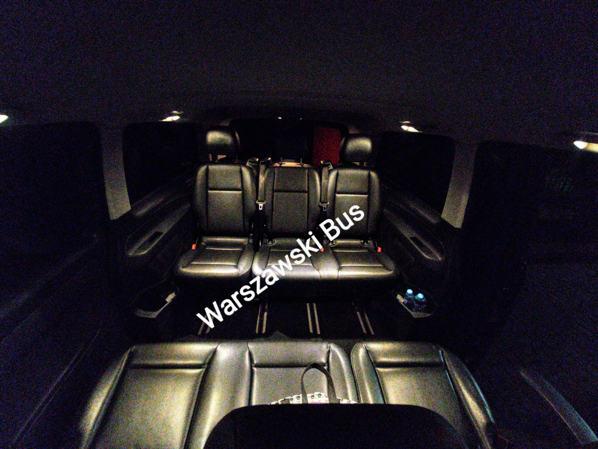 Wnętrze busa z trzema rzędami skórzanych, czarnych siedzeń, widok z tyłu pojazdu, słabe oświetlenie wnętrza.