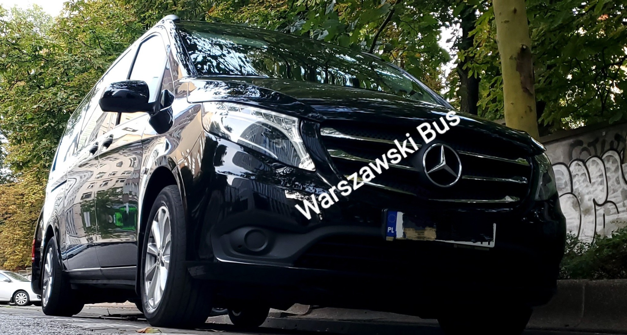 Czarny, lśniący Mercedes Vito z widoczną tablicą rejestracyjną PL, zaparkowany na tle zieleni miejskiej i muru z graffiti.