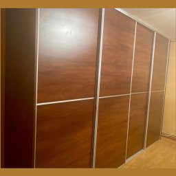 Serce Dębu - Duża szafa przesuwna z czterema drzwiami w odcieniach ciepłego brązu i srebrnymi listwami, widoczna z boku w jasnym pomieszczeniu.