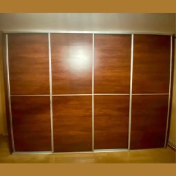 Serce Dębu - Duża szafa przesuwna z czterema drzwiami o ciemnym, drewnianym wykończeniu i srebrnych obramowaniach, wypełniająca całą ścianę.