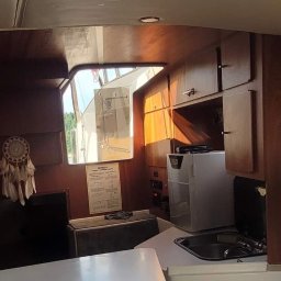 Serce Dębu - Wnętrze kabiny jachtu z drewnianą zabudową, widoczne szafki, zlewozmywak, mała lodówka, okno z widokiem na zewnątrz i łapacz snów na ścianie.