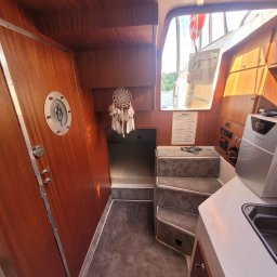 Serce Dębu - Wnętrze łodzi z drewnianą zabudową, schodkami z szarej wykładziny i widokiem na jezioro przez okno.