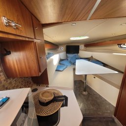 Serce Dębu - Wnętrze jachtu z drewnianymi szafkami, białą tapicerką, małym stolikiem i telewizorem. Na blacie kuchennym leży słomkowy kapelusz.