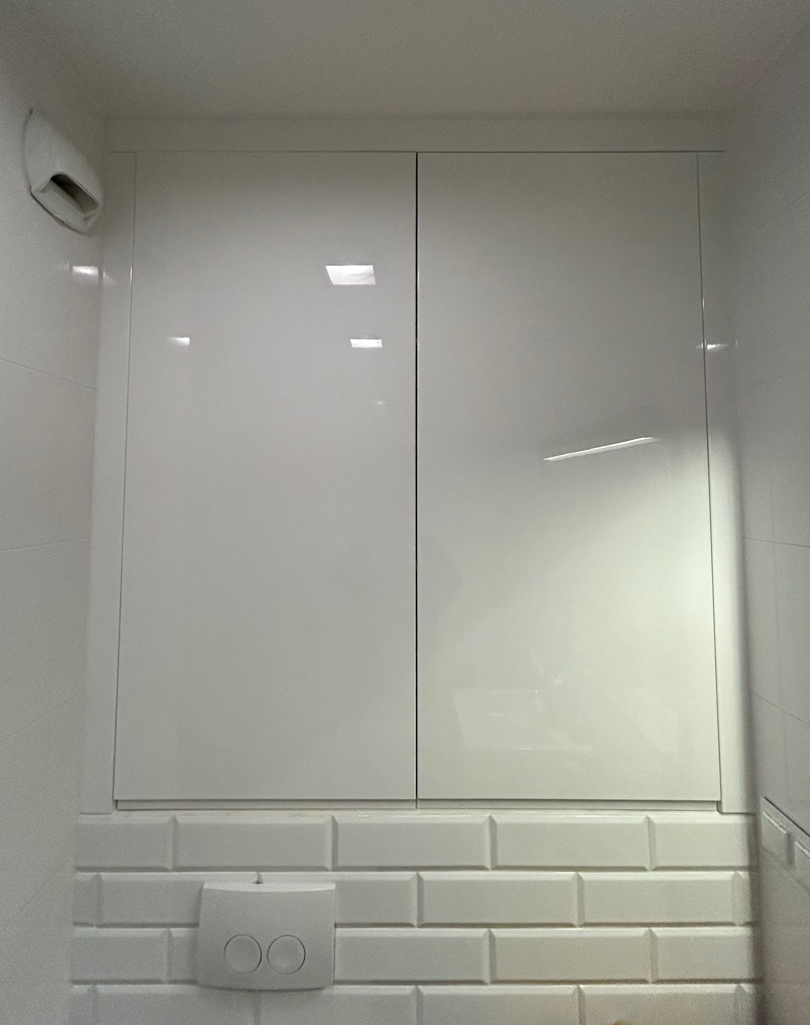 Biała, błyszcząca szafka w łazience, zamontowana nad spłuczką toaletową, z widocznymi płytkami ceramicznymi typu metro.