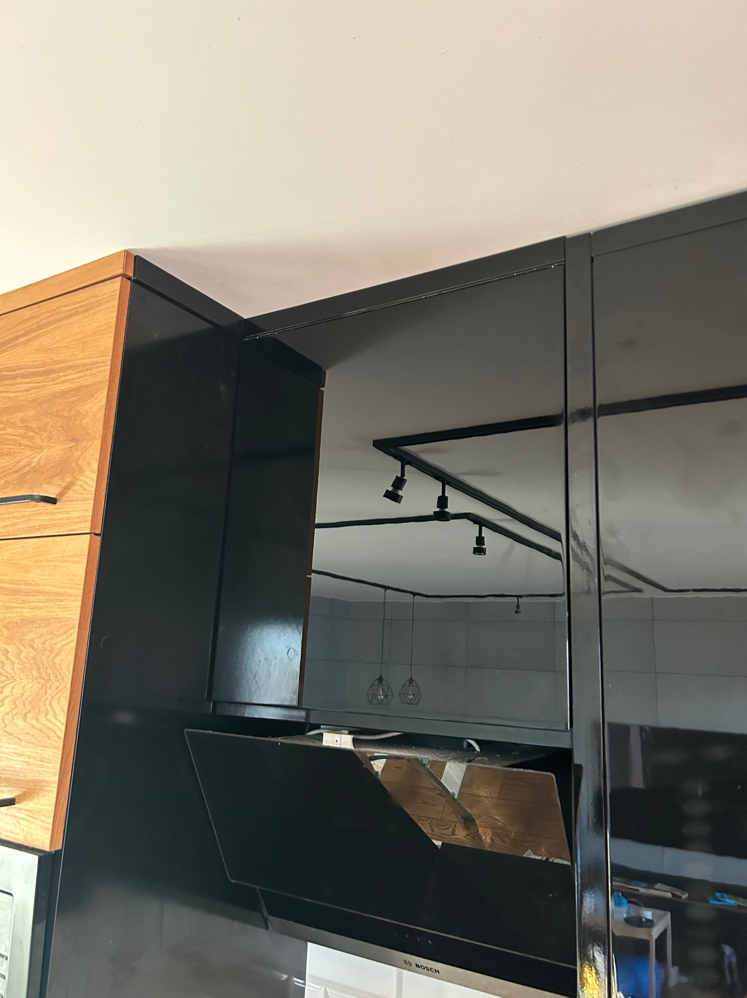Kątowe ujęcie czarnej szafki kuchennej z drewnianymi frontami, widoczny fragment okapu i odbicie oświetlenia sufitowego w lakierowanych powierzchniach.
