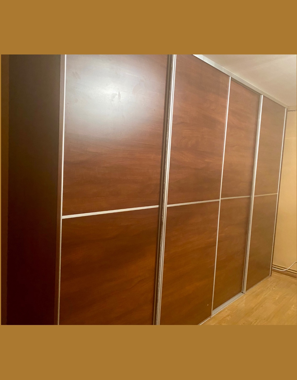 Duża szafa przesuwna z czterema drzwiami w odcieniach ciepłego brązu i srebrnymi listwami, widoczna z boku w jasnym pomieszczeniu.