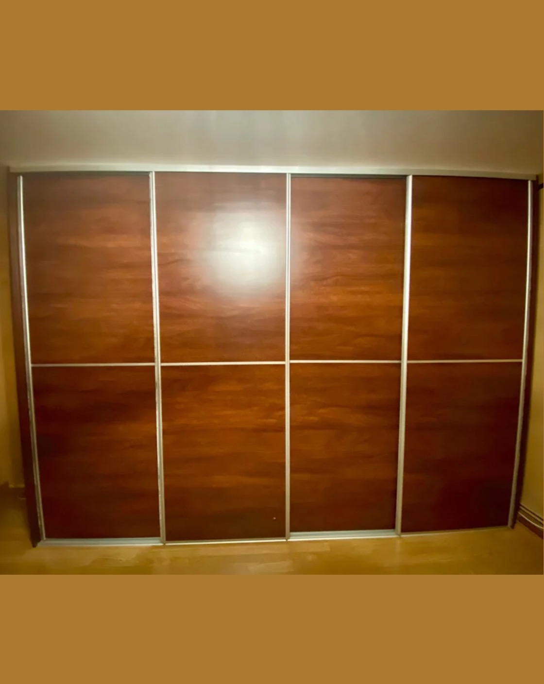 Duża szafa przesuwna z czterema drzwiami o ciemnym, drewnianym wykończeniu i srebrnych obramowaniach, wypełniająca całą ścianę.